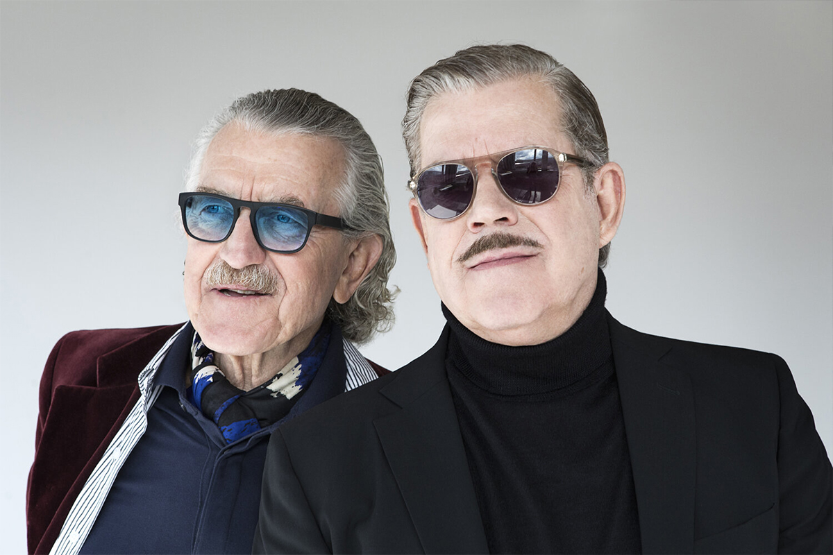 YELLO by Sobiralski at raw studios 03 A close-up portrait of Dieter Meier and Boris Blank, both wearing sunglasses and exuding effortless cool. Their contrasting outfits—one in a rich burgundy velvet blazer, the other in an all-black ensemble—capture their individual yet complementary styles.