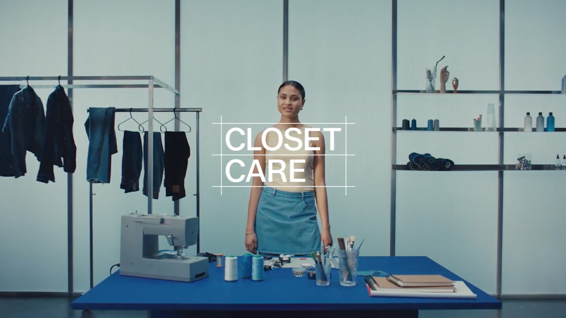 A clean, minimal studio scene featuring Alima Darouiche standing behind a table with sewing tools, denim garments, and a sewing machine. The words “CLOSET CARE” are overlaid in bold white text.