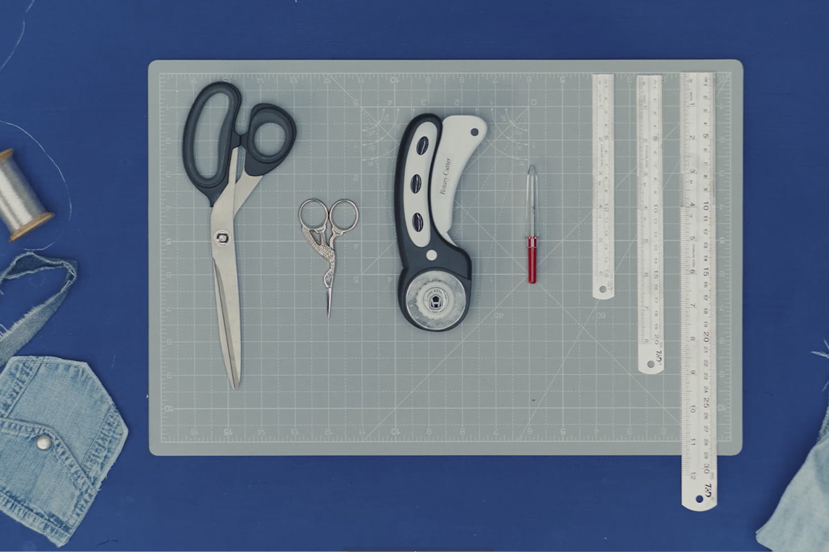 A top-down view of a cutting mat neatly arranged with sewing and measuring tools—scissors, rotary cutter, rulers, thread, and denim scraps—set on a vibrant blue background.