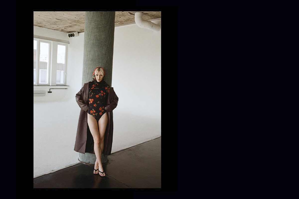 Luca Lorenz stands confidently in raw studios, leaning against a tiled pillar in a sunlit studio space, wearing a black floral bodysuit, long brown coat, and red headscarf from the Magda Butrym x H&M collection.