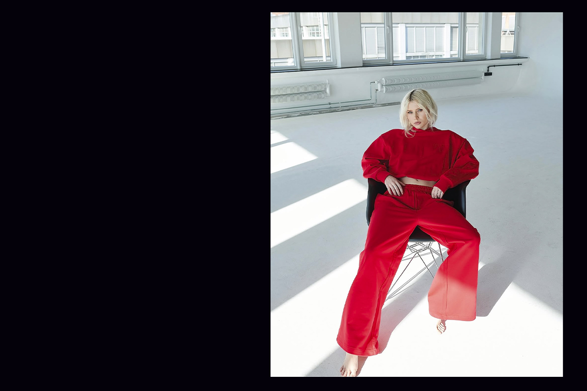 Luca Lorenz sits barefoot on a black chair in a bright, all-white studio, dressed in a bold red oversized sweatshirt and matching relaxed pants from the Magda Butrym x H&M collection.