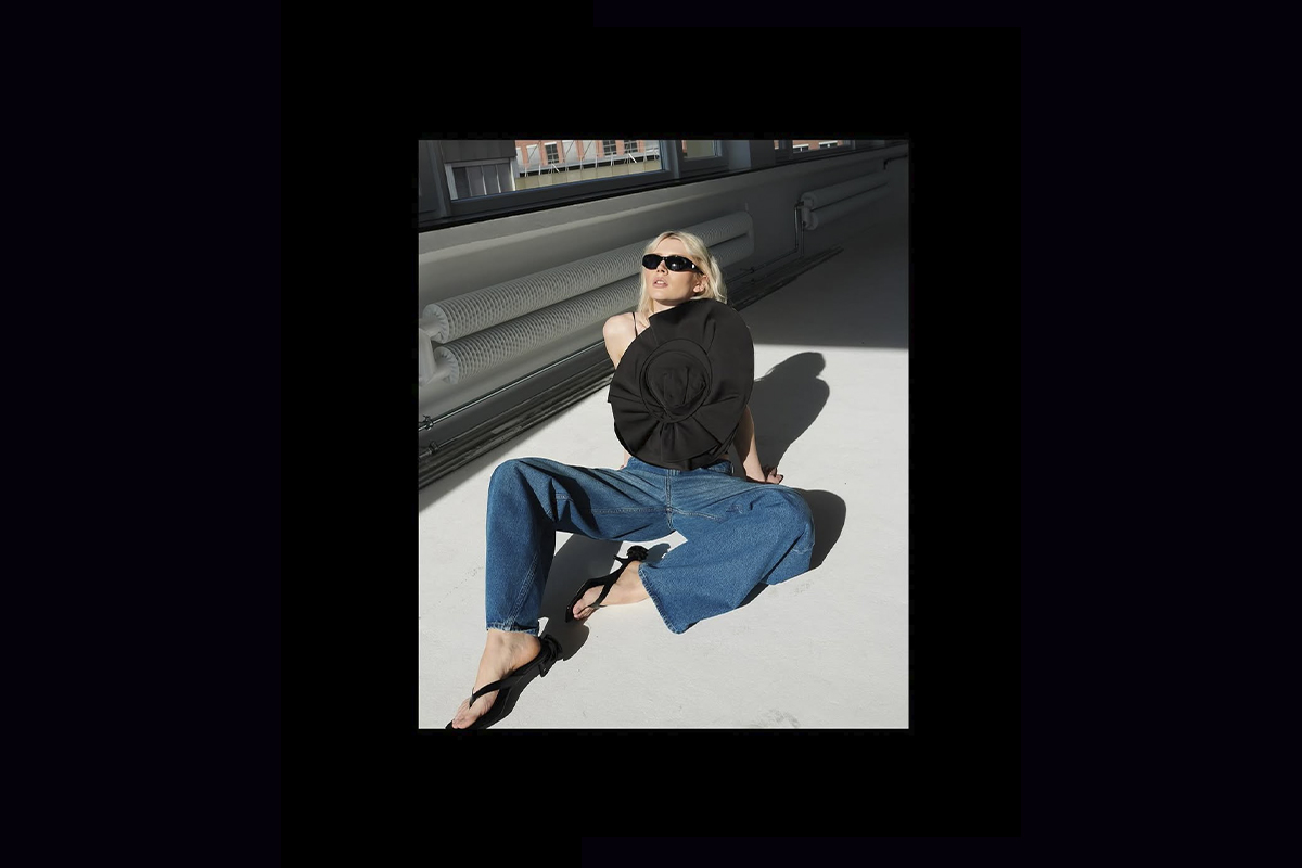 Luca Lorenz sits on the white studio floor with sunlight casting strong shadows, wearing blue wide-leg jeans, a sculptural black top with an oversized floral applique from the Magda Butrym x H&M collection.