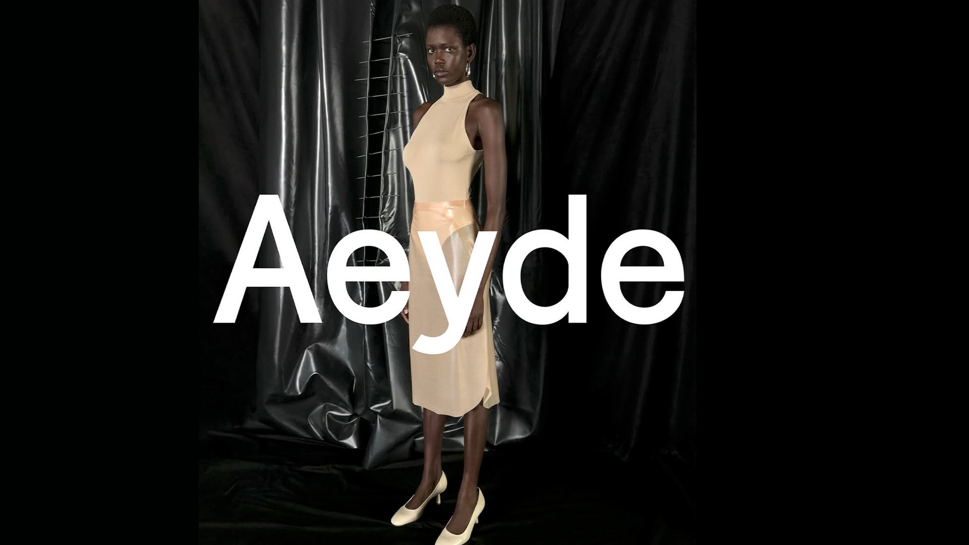Ajah Angau wearing a sheer beige dress and heels, standing against a glossy black backdrop with sculptural stitching elements – shot at raw studios for aeyde’s Pre-Spring 25 campaign.