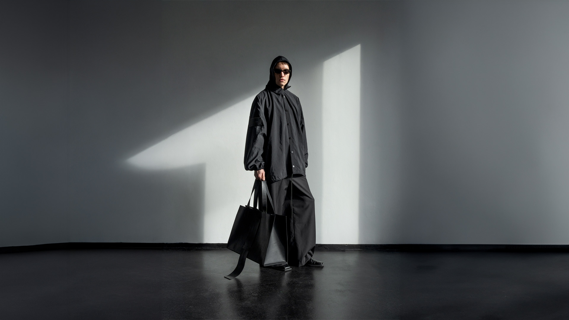 Model in an all-black oversized outfit and sunglasses stands in a patch of natural light on black concrete floor, against a grey wall at raw studios.