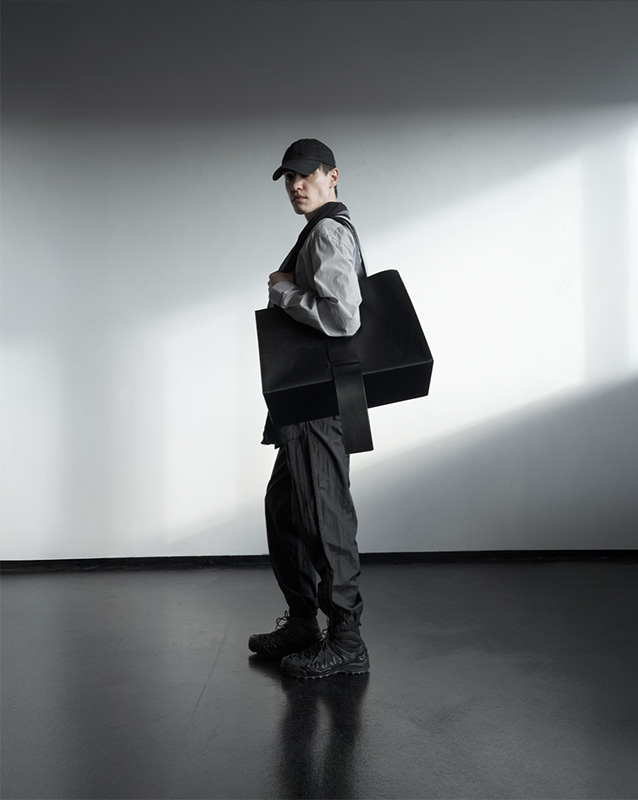 Model in a baseball cap and utility-style outfit carries a structured black leather tote, walking through a beam of natural light in Studio 2 at raw studios.