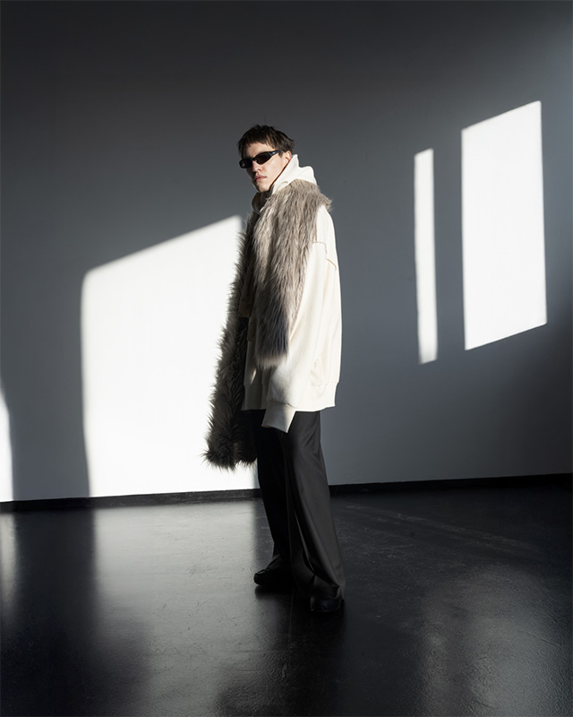 Model wearing a cream oversized hoodie and a dramatic faux-fur scarf poses in deep contrast lighting, framed by large rectangular window shadows at raw studios.